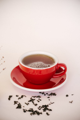A steaming hot tea in a red ceramic cup with tea leaves scattered around on a white background.