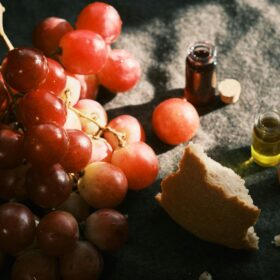 Elegant still life featuring grapes, broken bread, and small bottles on a dark fabric background.