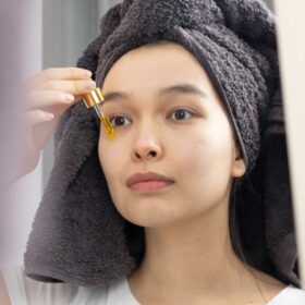 An Asian woman applies skincare serum with a dropper in front of a mirror.