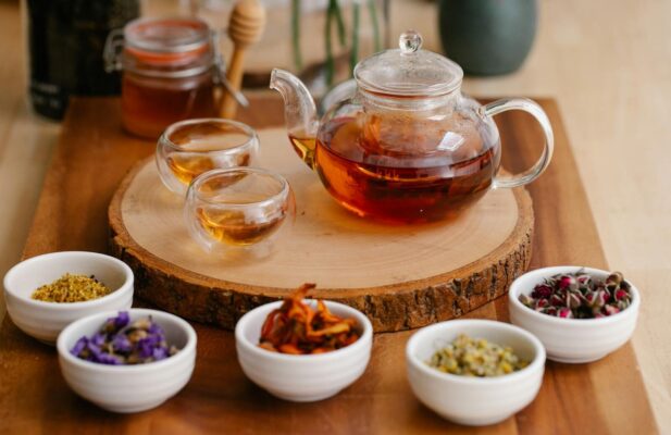 Glass teapot and cups with herbal infusions, fresh herbs, and honey dippers on a wooden tray.