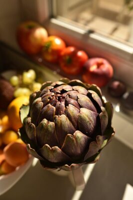 Close-up of a fresh artichoke surrounded by colorful fruits in a sunlit kitchen.