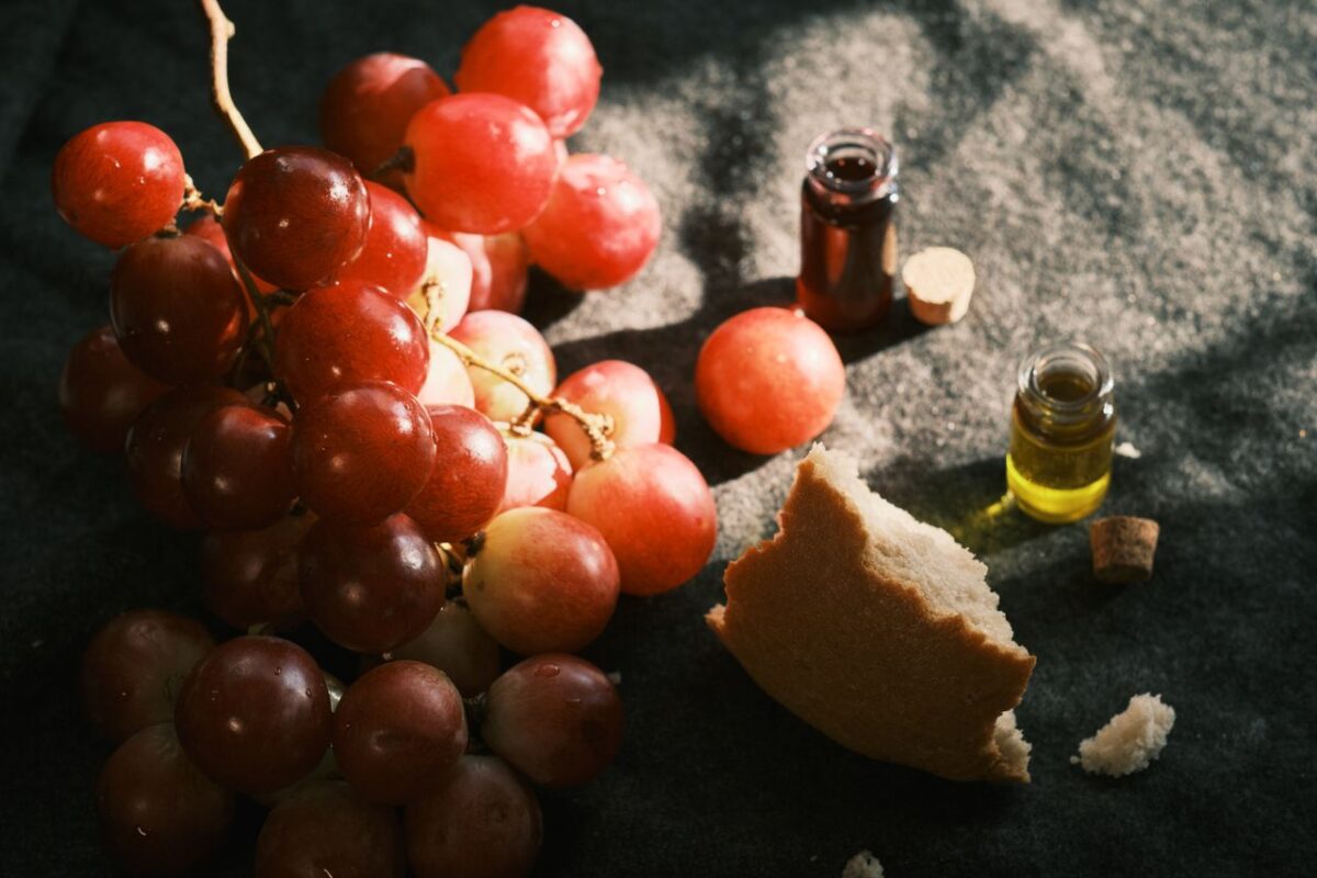 Elegant still life featuring grapes, broken bread, and small bottles on a dark fabric background.